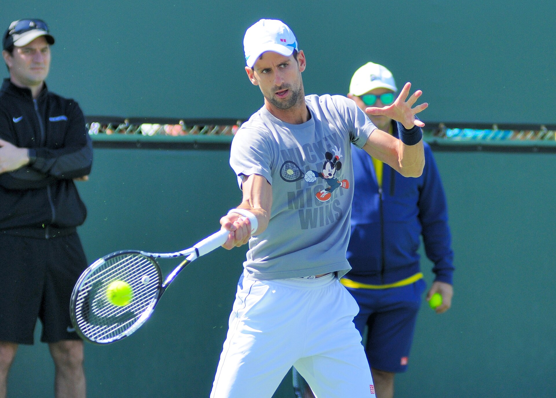 Novak Djokovic in a white cap and grey shirt making contact with a forehand.