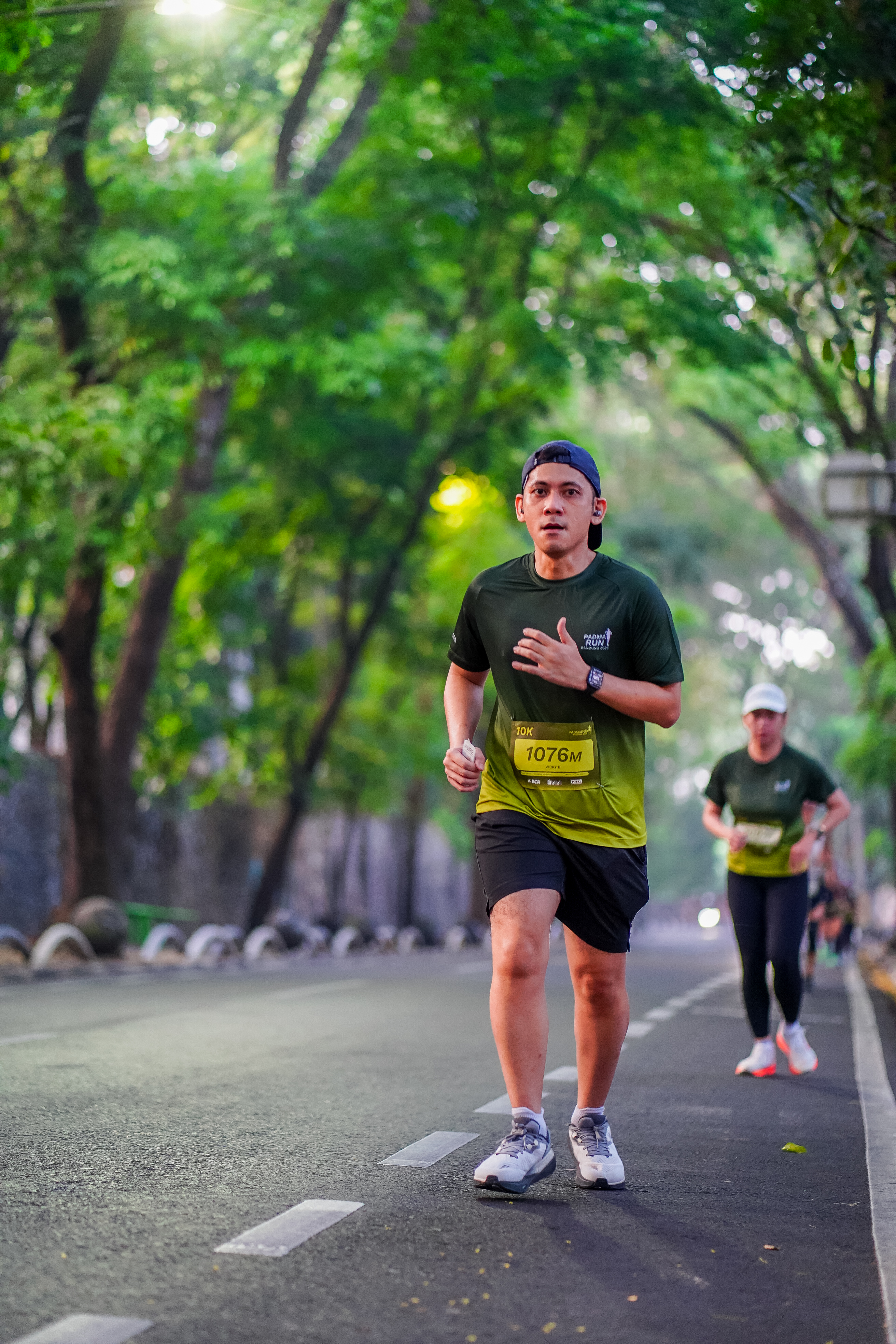 A runner in a green race shirt on a tree-lined road during the Padma Run 10K in Bandung.